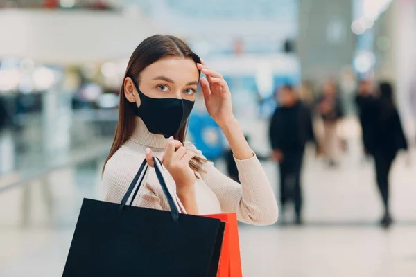 Mujer adulta joven que usa mascarilla médica protectora que lleva bolsas de papel en las manos en el centro comercial. Viernes Negro y covid-19 nuevo normal. —  Fotos de Stock