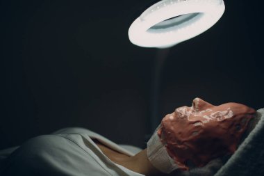 Yüz maskesi uygulama sürecini değiştir. Spatula tutan bir güzellik uzmanının dişi elleri. Kozmetik uzmanının ofisinde kanepede yatan bir kadın. Güzellik salonunda yüz bakımı.