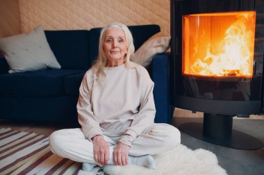 Oturma odasında şömineli koltukta oturan olgun gri saçlı kadın.