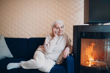 Yaşlı, gri saçlı kadın kanepede oturuyor ve şömineli akıllı telefonlardan konuşuyor.