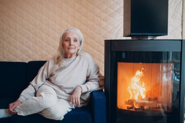 Oturma odasında şömineli koltukta oturan yaşlı gri saçlı kadın.