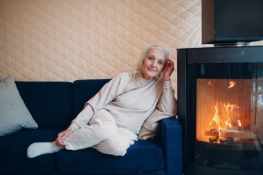 Oturma odasında şömineli koltukta oturan yaşlı gri saçlı kadın.