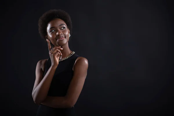 Jeune femme noire essayant de trouver la meilleure solution. Femme afro-américaine ayant une expression douteuse regardant du côté et tenant son menton avec la main sur fond noir. — Photo