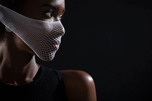 Retrato de primer plano de una mujer afroamericana modelo de moda que usa la máscara de la cara médica de cuarentena malla gruesa sobre fondo negro. Concepto de protección del coronavirus Covid-19. — Foto de Stock