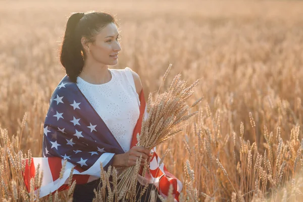 Donna con bandiera americana e con un covone di spighe nel campo di grano al tramonto. Il 4 luglio. Giorno di indipendenza vacanza patriottica. — Foto Stock