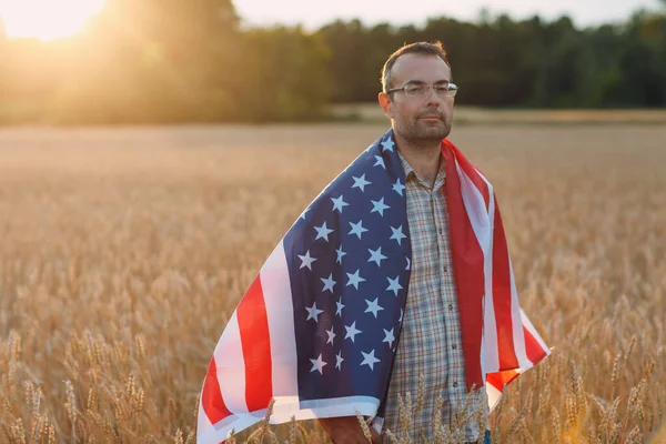 Ένας αγρότης που καλύπτει αμερικάνικη σημαία σε χωράφι με σιτάρι το ηλιοβασίλεμα. Γεωργία και γεωργική συγκομιδή, — Φωτογραφία Αρχείου