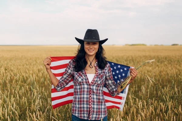 Femeie fermier purtând pălărie de cowboy, cămașă și blugi cu steag american pe câmpul de grâu — Fotografie, imagine de stoc