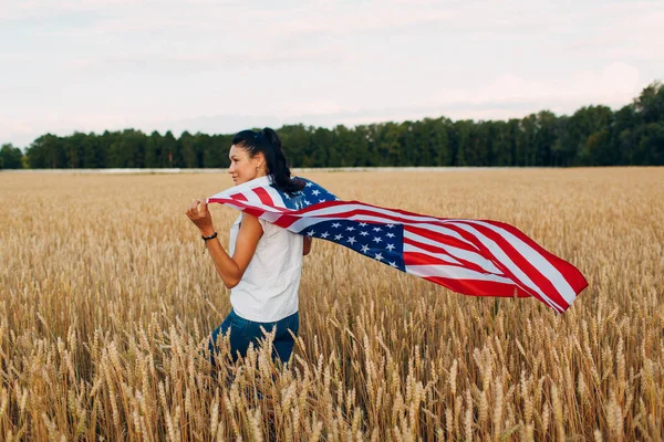 Mujer con bandera americana en el campo de trigo al atardecer. 4 de julio. Día de la Independencia fiesta patriótica. — Foto de Stock