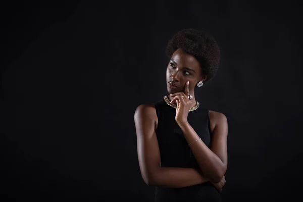 Young black woman trying to find best solution. African american female having doubtful expression looking side away and holding her chin with hand on black background.