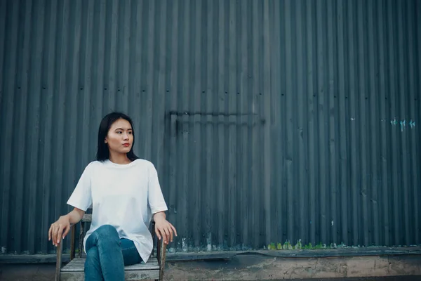 Urbano giovane donna asiatica seduta sulla sedia vicino muro blu sulla strada all'aperto. — Foto Stock