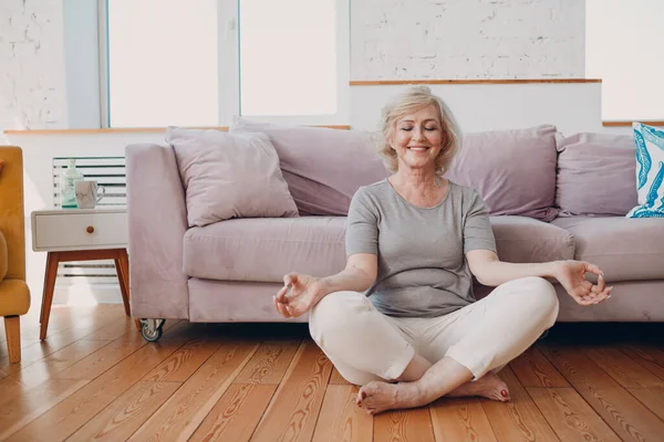 Oudere, lachende vrouw die thuis yoga beoefent. Ouderen ontspannen vrouw zitten in lotus poseren en mediteren zen als. — Stockfoto