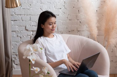 Asyalı kadın portresi ev içi oturma odasında dizüstü bilgisayar kullanmaktan zevk alıyor. Video çağrısı webinar akış konsepti.