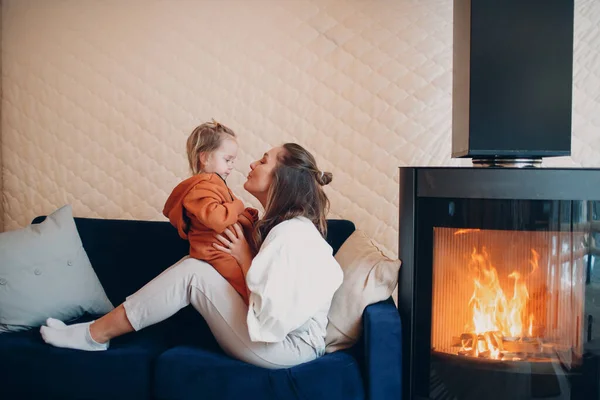 Moeder en kind zitten en spelen op de bank bij de open haard. Mam en baby. Ouder en kind ontspannen thuis. Familie hebben plezier samen. — Stockfoto