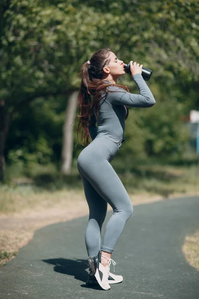 피곤 한 달리기 선수인 여자 가 공원 밖에서 조깅을 하다가 병에 든 생수를 마시고 있다. — 스톡 사진