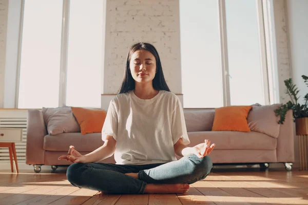 Aziatische vrouw doen yoga en zen als meditatie lotus poseren in casual dragen in een overdekte woonkamer appartement met natuurlijk zonlicht — Stockfoto