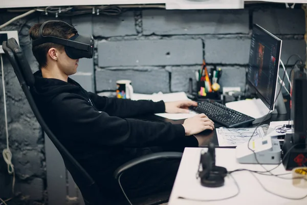 Joven programador gamer sentado en la silla con el escritorio de la computadora cerca de gafas de realidad virtual, gafas vr auriculares — Foto de Stock