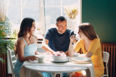 Erkek ve genç kadınlar evlerinin mutfağında, masada pastalarıyla oturup doğum günlerini kutluyorlar.