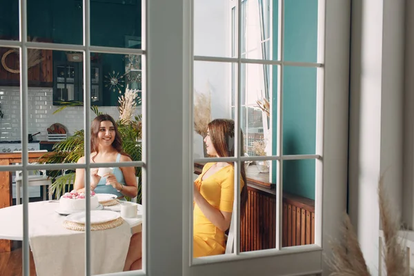 Perempuan senang teman-teman di rumah duduk dan tersenyum dengan kue ulang tahun putih di balik pintu kaca — Stok Foto