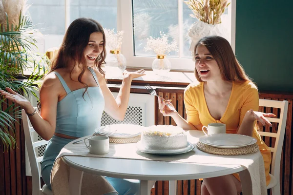 여자들은 집에 있는 행복 한 친구들 이 창가에 앉아 하얀 생일 케이크와 함께 웃는 모습을 즐긴다 — 스톡 사진