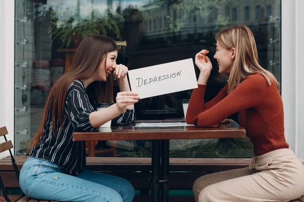 Concetto di depressione nascosta. Donna che tiene in mano un foglio di carta bianca con scritto Depressione. Due donne che parlano e sorridono nel caffè di strada. — Foto Stock