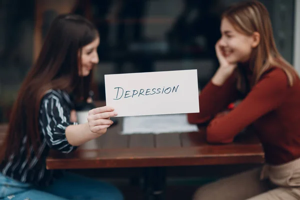 Versteckte Depression. Frau mit weißem Blatt Papier mit der Aufschrift Depression in der Hand. Zwei Frauen reden und lächeln in Straßencafé. — Stockfoto
