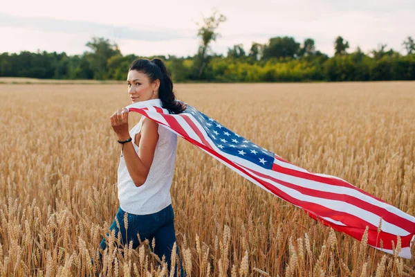 Mujer con bandera americana en el campo de trigo al atardecer. 4 de julio. Día de la Independencia fiesta patriótica. — Foto de Stock