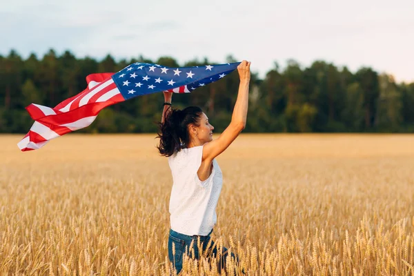 Femeie cu steag american în câmp de grâu la apusul soarelui. 4 iulie. Ziua Independenței sărbătoare patriotică . — Fotografie, imagine de stoc