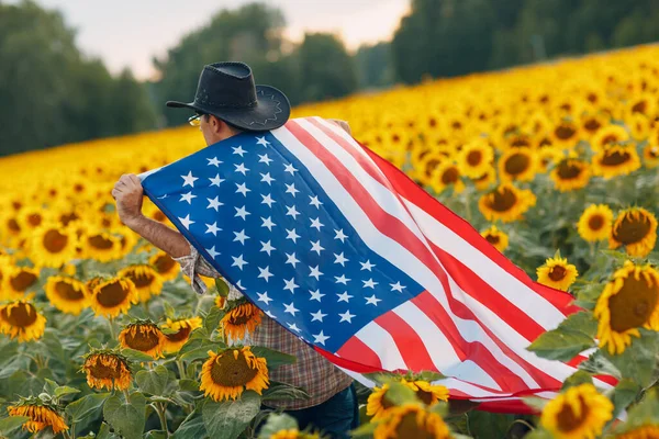 Ένας άντρας μπαίνει στο χωράφι με τα ηλιοτρόπια με τη σημαία των Ηνωμένων Πολιτειών. 4η Ιουλίου Έννοια Ημέρα Ανεξαρτησίας. — Φωτογραφία Αρχείου