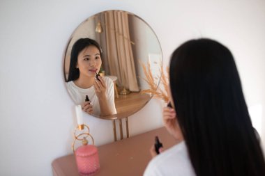 Aynanın önündeki güzel Asyalı kadın oturma odasında ruj sürerek makyaj yapıyor.