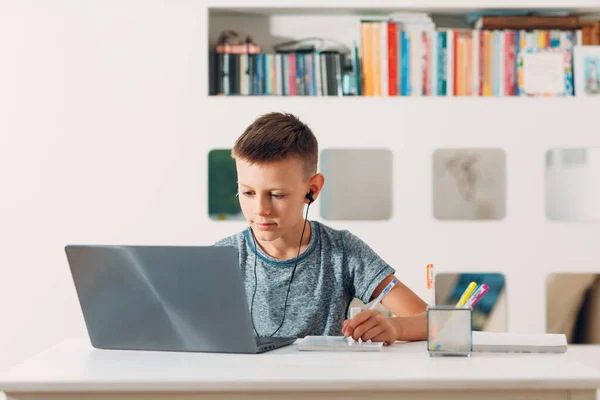Młody chłopak w słuchawkach siedzi przy stole z laptopem i przygotowuje się do szkoły. Koncepcja edukacji internetowej. — Zdjęcie stockowe