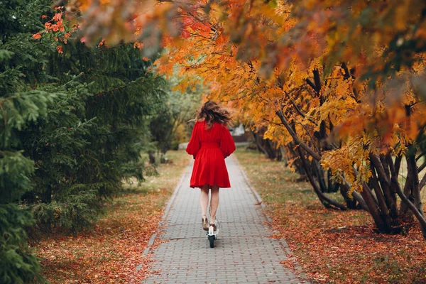 Young woman with electric scooter in red dress at the autumn city park — Stock Photo, Image