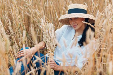 Hasır şapkalı genç kadın tarlada buğday kulakları tutuyor.