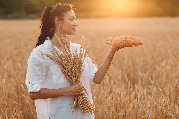 Donna che tiene il pane di grano fatto in casa e il covone di orecchie nelle mani nel campo di grano al tramonto. — Foto Stock