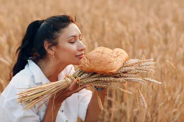 Donna che tiene e annusa Pane di grano fatto in casa e covone di orecchie nelle mani nel campo di grano al tramonto. — Foto Stock