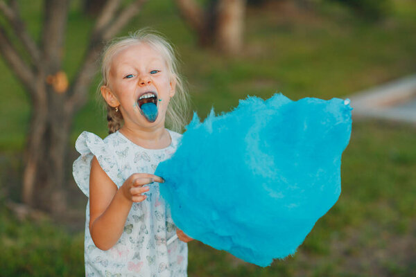 Маленькая блондинка ест сладкую вату и показывает синий язык в парке.