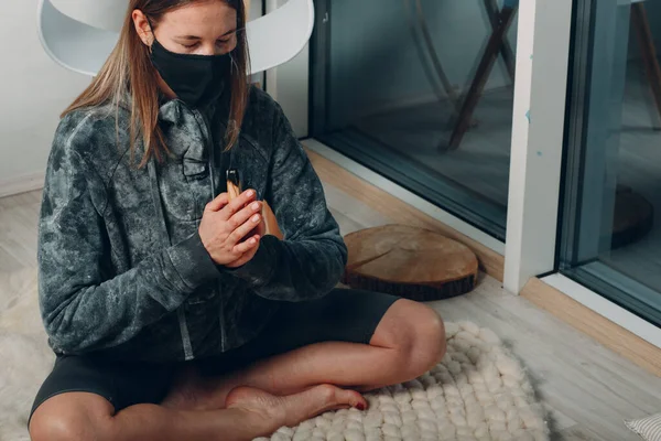 Volwassen volwassen vrouw doet yoga in het gezicht masker thuis woonkamer met roken palo santo stok boom — Stockfoto