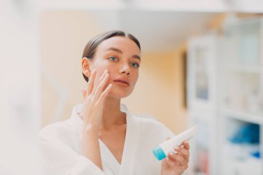 Genç bir kadın yüzüne kozmetik beyaz krem sürüyor.