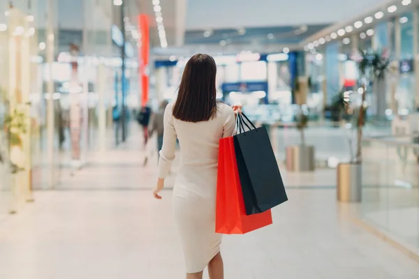 Mujer adulta joven positiva que lleva bolsas de papel en las manos. Vista trasera trasera —  Fotos de Stock