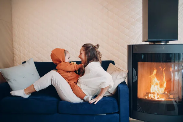 Madre e hijo sentados y jugando en el sofá cerca de la chimenea. Mamá y el bebé. Padre e hijo pequeño relajándose en casa. Familia divirtiéndose juntos. — Foto de Stock