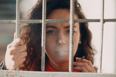 Hapishane parmaklıklarının arkasında sigara içen turuncu takım elbiseli, esmer, kıvırcık kadın. Renkli tulumlu bir kadın portresi..