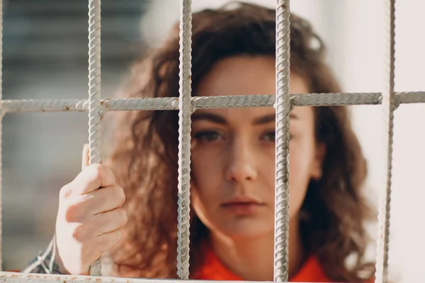 Jovem morena encaracolada em terno laranja atrás das grades da prisão. Feminino em macacão colorido retrato. — Fotografia de Stock