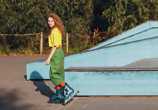 Jonge vrouw meisje in groene en gele kleding en oranje kousen met krullend kapsel rolschaatsen in skate park — Stockfoto