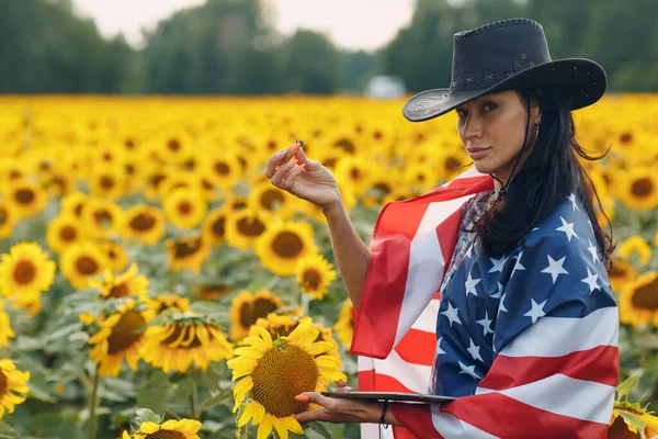 ひまわり畑にアメリカ国旗とタブレットPCを持つ若い女性農家 — ストック写真