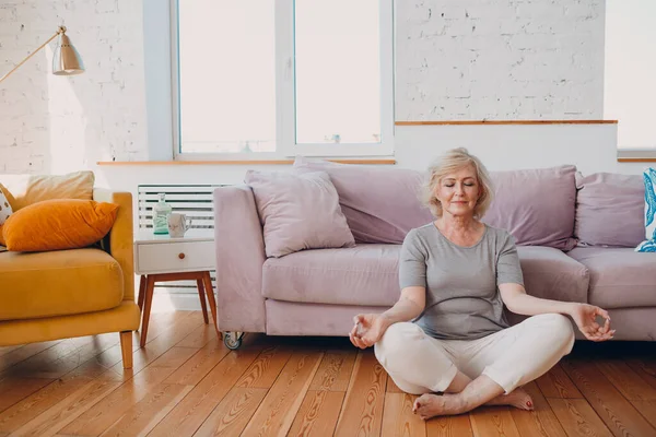 Oudere, lachende vrouw die thuis yoga beoefent. Ouderen ontspannen vrouw zitten in lotus poseren en mediteren zen als. — Stockfoto