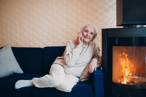 Wanita tua berambut abu-abu duduk di sofa dan berbicara smartphone di ruang tamu dengan perapian — Stok Foto