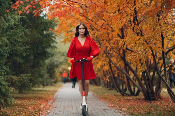 Jovem com scooter elétrico em vestido vermelho no parque da cidade de outono — Fotografia de Stock