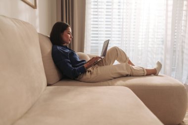 Bir kadın rahat bir şekilde kanepede oturur, dizüstü bilgisayarını kullanır ve doğal ışığın tadını çıkarır.