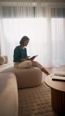 Kadın, güneşli oturma odasında tablet okuyor, perdeleri dağınık, modern poziyonlu.