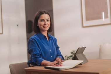 Genç bir kadın ofis masasında çalışırken elinde tablet ve defterle gülümsüyor..