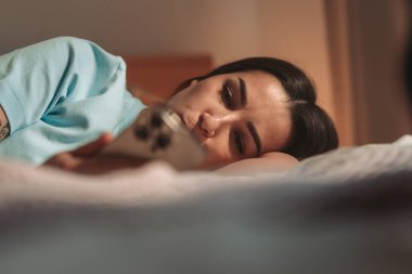 Yatakta uzanmış genç bir kadın telefon yemliği, loş bir ışık, rahat bir duruş, hoş bir gülümseme, akşam vakti.